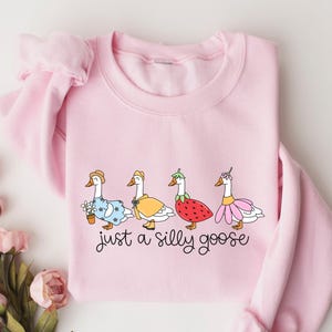 Peut inclure: Un sweat-shirt rose clair avec une illustration fantaisiste de quatre oies v&ecirc;tues de tenues vari&eacute;es. Les oies sont orn&eacute;es de chapeaux, de robes et d'accessoires. Le texte "just a silly goose" est &eacute;crit dans une police ludique en dessous.