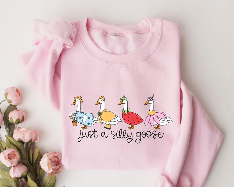 Peut inclure: Un sweat-shirt rose clair avec une illustration fantaisiste de quatre oies v&ecirc;tues de tenues vari&eacute;es. Les oies sont orn&eacute;es de chapeaux, de robes et d'accessoires. Le texte "just a silly goose" est &eacute;crit dans une police ludique en dessous.