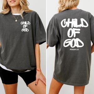 May include: Dark gray t-shirt with white text that reads "CHILD OF GOD" on the front and back. The back of the shirt also includes "PSALM 23" in white text. The shirt has a relaxed fit and short sleeves.