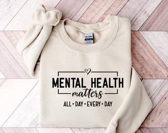 May include: A cream-colored sweatshirt with the words "MENTAL HEALTH matters" in black, framed by a rectangle and a heart. Below the text are the words "ALL • DAY • EVERY • DAY". The sweatshirt is folded on a wooden surface.