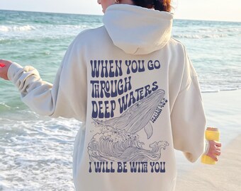 May include: Cream-colored hoodie with a whale graphic and the text "WHEN YOU GO THROUGH DEEP WATERS I WILL BE WITH YOU" in navy blue. The hoodie has a hood and long sleeves, suitable for casual wear.