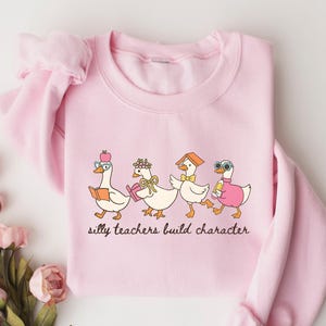 May include: A light pink sweatshirt featuring a whimsical illustration of four cartoon geese, each adorned with accessories like glasses and hats. The geese are walking in a line, and the text "silly teachers build character" is printed below the geese.