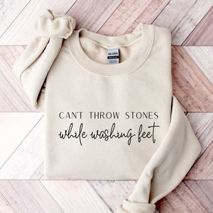 May include: A beige sweatshirt with the text "Can't throw stones while washing feet" printed on the front.
