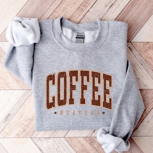 Puede incluir: Sudadera gris con texto marr&oacute;n que dice "COFFEE WEATHER" con una estrella a cada lado del texto.