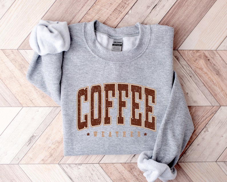 Puede incluir: Sudadera gris con texto marr&oacute;n que dice "COFFEE WEATHER" con una estrella a cada lado del texto.