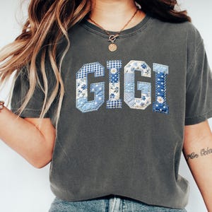 May include: A dark gray t-shirt with the word "GIGI" in blue patchwork letters. The letters feature floral, gingham, and polka dot patterns. The shirt has a crew neck and short sleeves.
