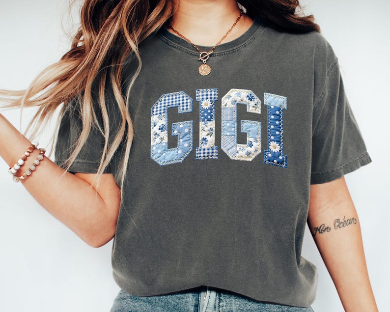 May include: A dark gray t-shirt with the word "GIGI" in blue patchwork letters. The letters feature floral, gingham, and polka dot patterns. The shirt has a crew neck and short sleeves.