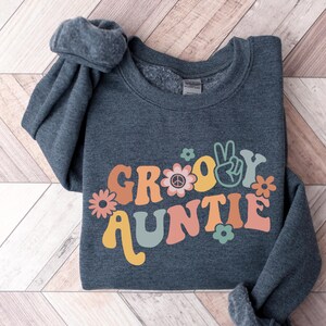 May include: A dark gray sweatshirt with the text "Groovy Auntie" in colorful retro lettering. The text is decorated with peace signs and flowers.