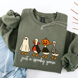 May include: An olive green sweatshirt with the phrase "just a spooky goose" in white script. The graphic features four cartoon geese dressed as a ghost, vampire, jack-o'-lantern, and skeleton. A silver necklace is draped across the sweatshirt.