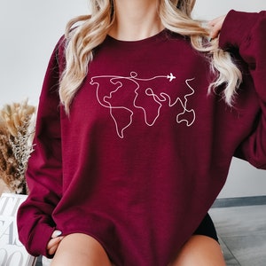 Op de afbeelding: Een bordeauxrood sweatshirt met een witte omtrek van een wereldkaart en een klein vliegtuig dat erboven vliegt.