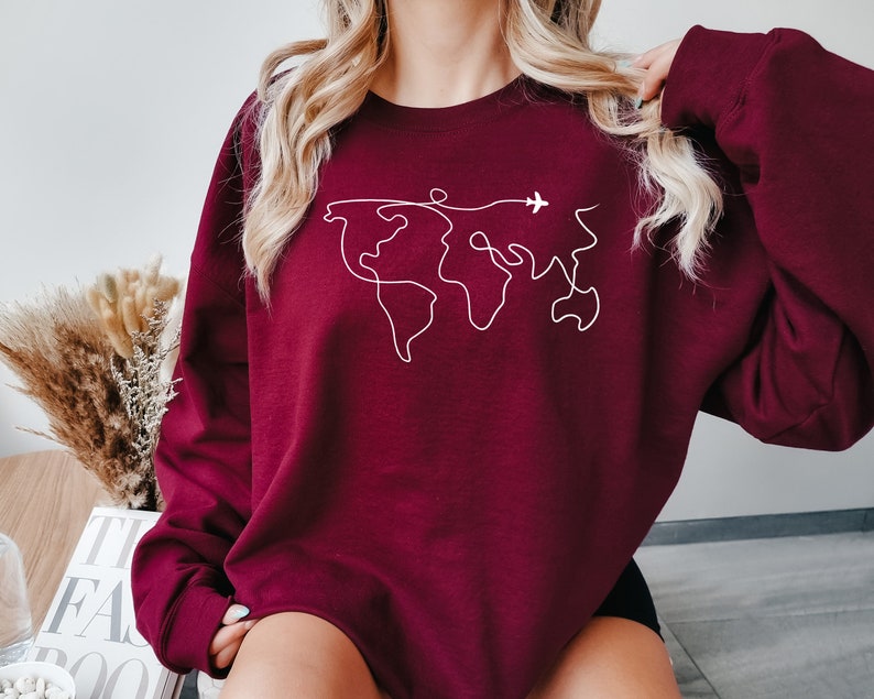 Op de afbeelding: Een bordeauxrood sweatshirt met een witte omtrek van een wereldkaart en een klein vliegtuig dat erboven vliegt.