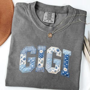 May include: Gray Comfort Colors t-shirt with the name "GIGI" appliqued in blue and white patchwork fabric. The letters feature floral, polka dot, and gingham patterns. A gold necklace with a pendant rests on the shirt.