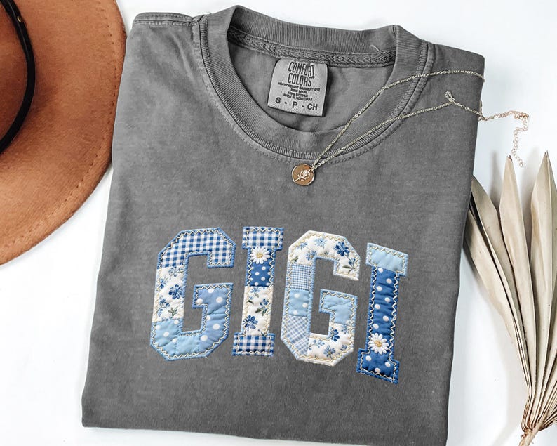 May include: Gray Comfort Colors t-shirt with the name "GIGI" appliqued in blue and white patchwork fabric. The letters feature floral, polka dot, and gingham patterns. A gold necklace with a pendant rests on the shirt.