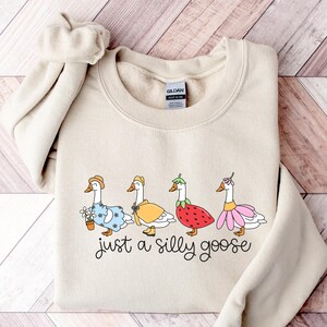 Peut inclure: Sweat-shirt beige avec une illustration fantaisiste de quatre oies v&ecirc;tues de tenues vari&eacute;es, dont un chapeau, une cape et une robe &agrave; fleurs. Le texte "just a silly goose" est &eacute;crit en dessous des oies dans une police manuscrite.