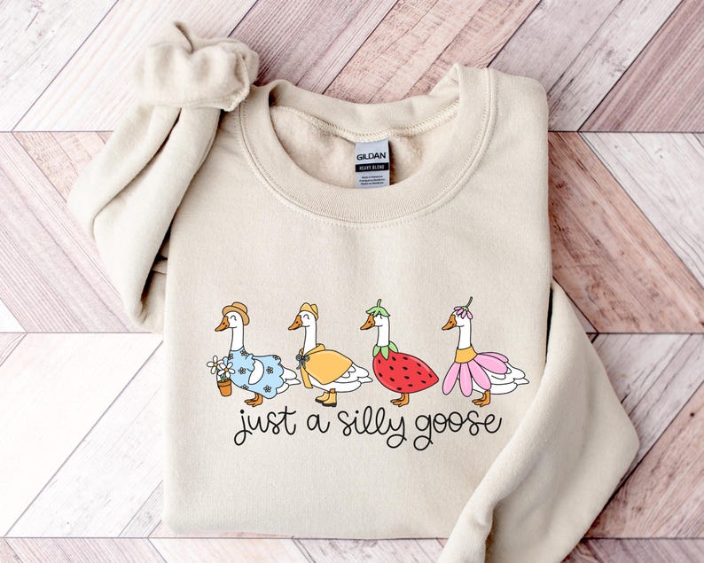 Peut inclure: Sweat-shirt beige avec une illustration fantaisiste de quatre oies v&ecirc;tues de tenues vari&eacute;es, dont un chapeau, une cape et une robe &agrave; fleurs. Le texte "just a silly goose" est &eacute;crit en dessous des oies dans une police manuscrite.