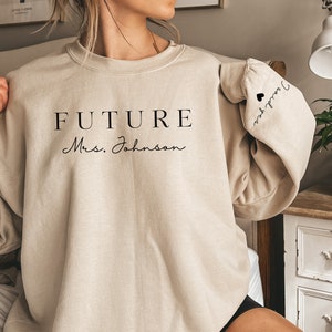 May include: Beige sweatshirt with the text "FUTURE Mrs. Johnson" on the front and "I said yes" on the sleeve.