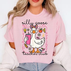 May include: A light pink t-shirt with a cartoon goose design. The goose holds a pink drink and is surrounded by school-themed graphics. The text "silly goose teacher club" is above and below the goose illustration.