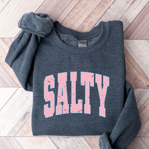May include: A dark gray sweatshirt with the word "SALTY" printed in pink letters. The letters have a distressed, vintage look.