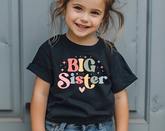 Op de afbeelding: Een zwart T-shirt met de tekst "BIG Sister" in kleurrijke letters met regenboogaccenten en kleine sterren.