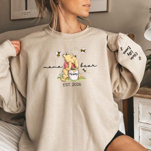 May include: Beige sweatshirt featuring a Winnie the Pooh graphic with the text "mama bear" and "Hunny". The design includes bees and the text "EST. 2026". The sleeve has the names "Olivia, Emma, Lilly" and bee graphics.