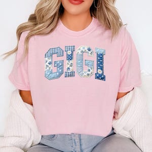 May include: A light pink t-shirt with the name "GIGI" in blue patchwork letters. The letters feature various blue floral and patterned fabrics. The shirt is a casual, crew neck style.
