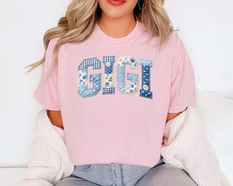 May include: A light pink t-shirt with the name "GIGI" in blue patchwork letters. The letters feature various blue floral and patterned fabrics. The shirt is a casual, crew neck style.
