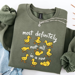 May include: An olive green sweatshirt with the phrase "most definitely not all in a row" and cartoon ducks. The ducks are yellow and the text is white. A silver necklace is draped over the sweatshirt.