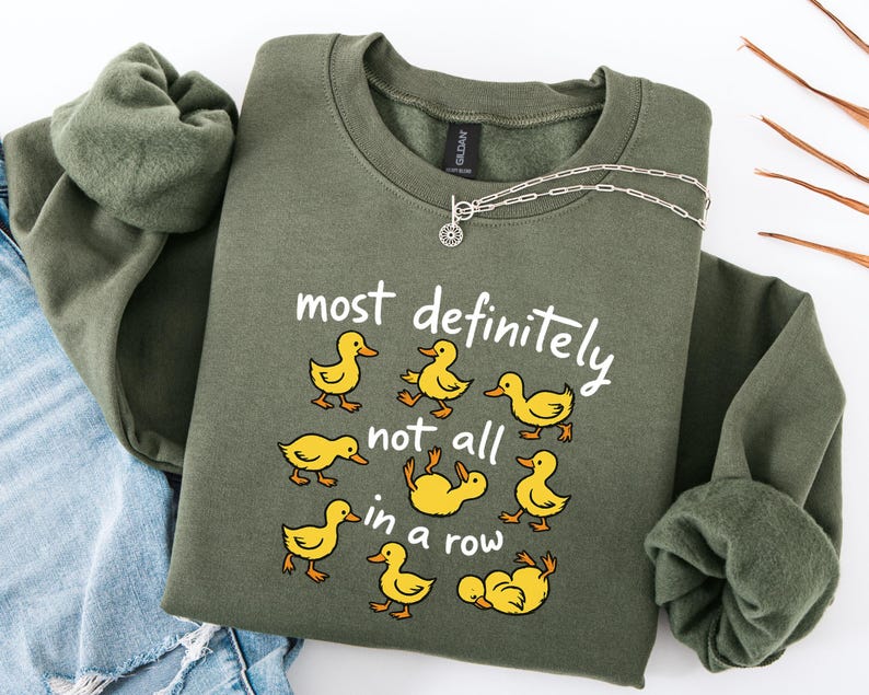 May include: An olive green sweatshirt with the phrase "most definitely not all in a row" and cartoon ducks. The ducks are yellow and the text is white. A silver necklace is draped over the sweatshirt.