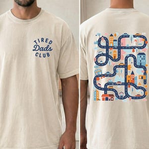 May include: Cream-colored t-shirt with the text "Tired Dads Club" in blue on the front. The back features a colorful graphic of a road map with cars, buildings, and trees. A casual, comfortable tee.