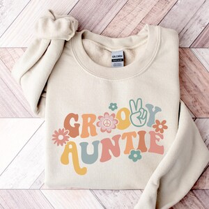 May include: A beige sweatshirt with the text "Groovy Auntie" in a retro font with peace signs and flowers.