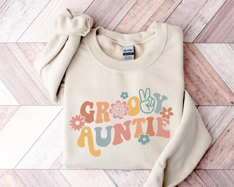 May include: A beige sweatshirt with the text "Groovy Auntie" in a retro font with peace signs and flowers.