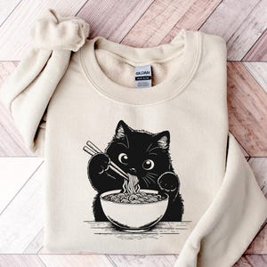 May include: Cream-colored sweatshirt featuring a black and white graphic of a cat eating noodles with chopsticks from a bowl. The sweatshirt has a crew neck and long sleeves. The brand tag is visible on the upper back.