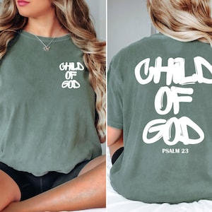 May include: Sage green t-shirt with the text "CHILD OF GOD" in white on the front and back. The back of the shirt also includes "PSALM 23" in white. The shirt is a short-sleeved, crew neck style.