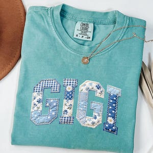 May include: A turquoise Comfort Colors t-shirt with the name "GIGI" appliqued in patchwork fabric. The letters feature floral, gingham, and polka dot patterns in shades of blue and white. A gold necklace with a pendant is draped across the shirt.