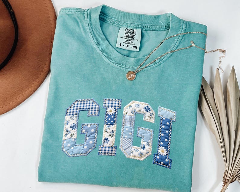 May include: A turquoise Comfort Colors t-shirt with the name "GIGI" appliqued in patchwork fabric. The letters feature floral, gingham, and polka dot patterns in shades of blue and white. A gold necklace with a pendant is draped across the shirt.