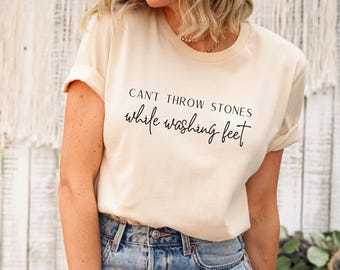 May include: Cream-colored t-shirt with the text "CAN'T THROW STONES while washing feet" in black script. The shirt has rolled-up sleeves and is being worn by a person. The shirt is paired with blue jeans.