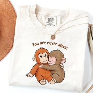 May include: Cream-colored t-shirt featuring a cartoon illustration of two hugging monkeys. The text "You are never alone" is printed above the monkeys in brown. A gold necklace and a brown hat are also visible.