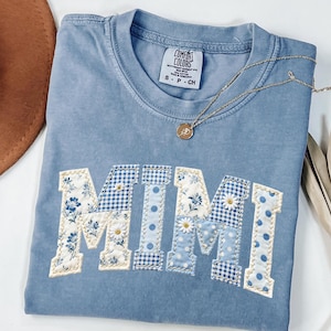 K&ouml;nnte beinhalten: Hellblaues Comfort Colors T-Shirt mit dem Wort "MIMI" auf der Vorderseite. Die Buchstaben bestehen aus verschiedenen blau-wei&szlig; gemusterten Stoffen, darunter Blumen-, Vichy- und Tupfenmuster. Eine goldene Halskette mit Anh&auml;nger liegt auf dem Shirt.