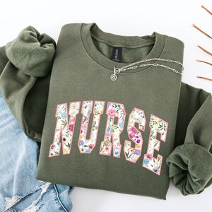 May include: A green sweatshirt with a floral print design that says "NURSE" in a white outline. The sweatshirt has a crew neck and long sleeves.