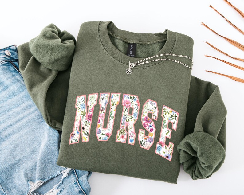 May include: A green sweatshirt with a floral print design that says "NURSE" in a white outline. The sweatshirt has a crew neck and long sleeves.