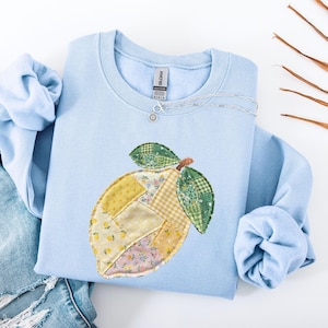 May include: A light blue sweatshirt with a patchwork lemon design. The lemon features various fabric patterns in yellow, green, and floral prints. A silver necklace with a small charm rests on the shirt. The sleeves are folded, and a pair of blue jeans is visible.