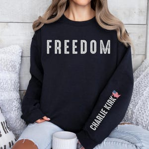 May include: A navy blue sweatshirt with the word "FREEDOM" printed across the chest in distressed white lettering. The right sleeve features the text "CHARLIE KIRK" in white, with a small American flag graphic.