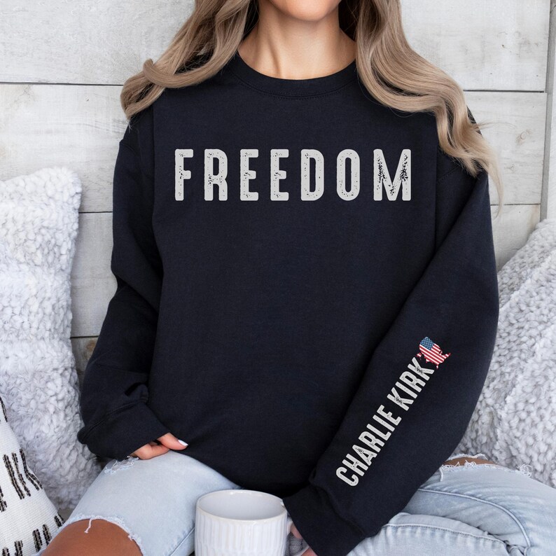 May include: A navy blue sweatshirt with the word "FREEDOM" printed across the chest in distressed white lettering. The right sleeve features the text "CHARLIE KIRK" in white, with a small American flag graphic.