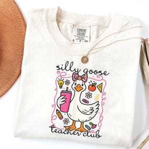 May include: Cream-colored t-shirt featuring a cartoon goose with a bow, holding a pink tumbler. The shirt has the text "silly goose teacher club" in black script, with colorful school-themed graphics.