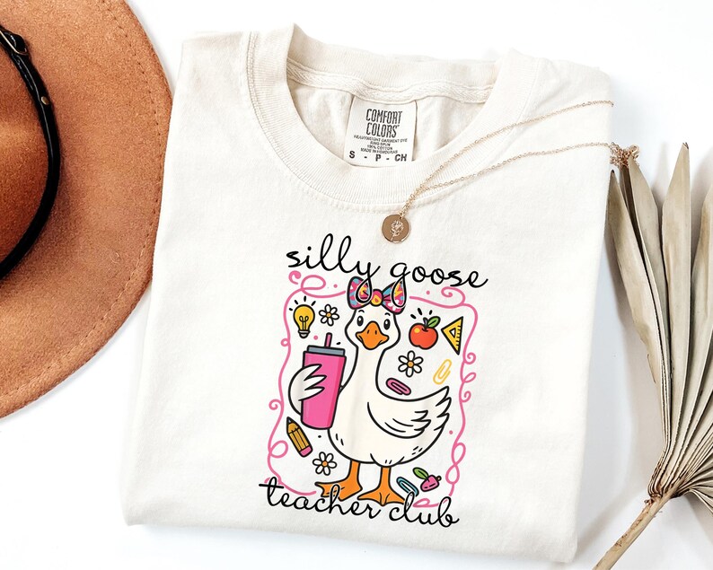 May include: Cream-colored t-shirt featuring a cartoon goose with a bow, holding a pink tumbler. The shirt has the text "silly goose teacher club" in black script, with colorful school-themed graphics.