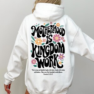 May include: White hoodie with a black graphic print that says "Motherhood is Kingdom Work" with floral accents. The text is in a retro font style. The quote "She is strong and dignified, laughs at the future. Speaks with wisdom and kindness. Takes care of her household, avoids idleness. Proverbs 31:25-27" is printed below the graphic.