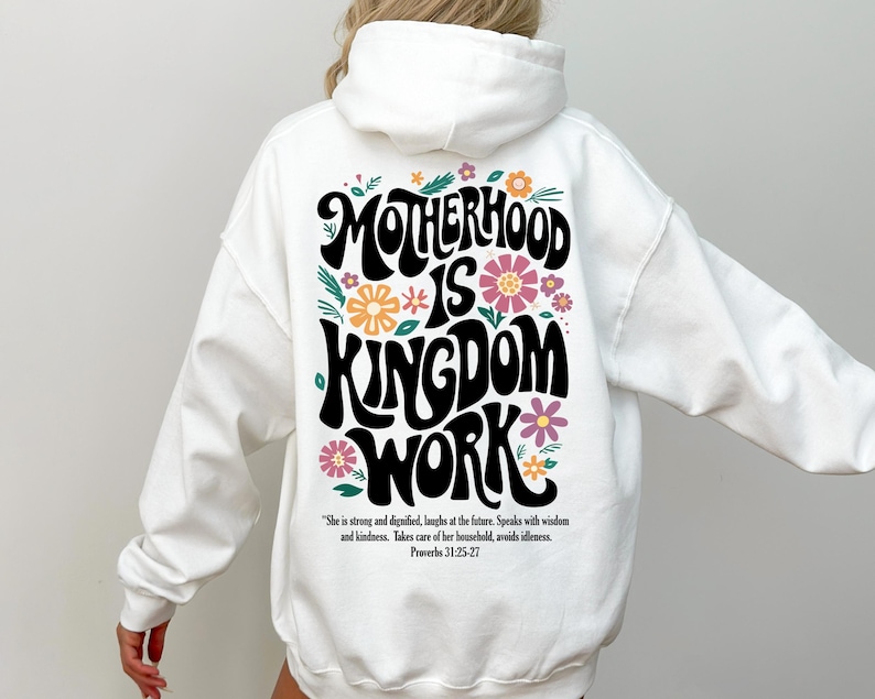 May include: White hoodie with a black graphic print that says "Motherhood is Kingdom Work" with floral accents. The text is in a retro font style. The quote "She is strong and dignified, laughs at the future. Speaks with wisdom and kindness. Takes care of her household, avoids idleness. Proverbs 31:25-27" is printed below the graphic.