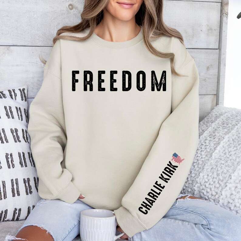 May include: Cream-colored sweatshirt with the word "FREEDOM" printed in large, black, distressed letters across the chest. The right sleeve features the text "CHARLIE KIRK" and a small American flag graphic.