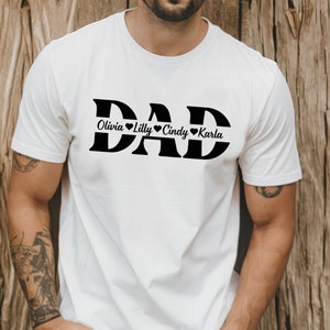 May include: White t-shirt with the word "DAD" in large black letters. The names "Olivia", "Lilly", "Cindy", and "Karla" are written in smaller black letters below the word "DAD" with a heart symbol between each name.
