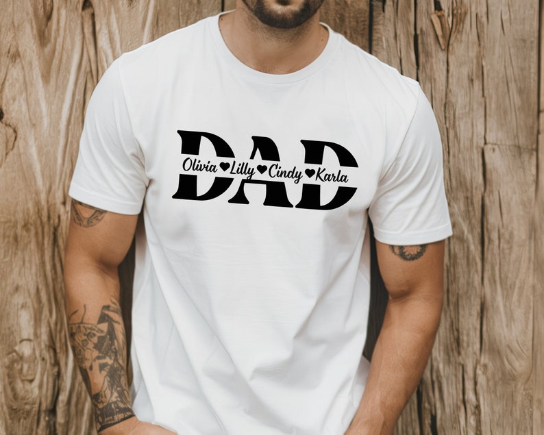 May include: White t-shirt with the word "DAD" in large black letters. The names "Olivia", "Lilly", "Cindy", and "Karla" are written in smaller black letters below the word "DAD" with a heart symbol between each name.